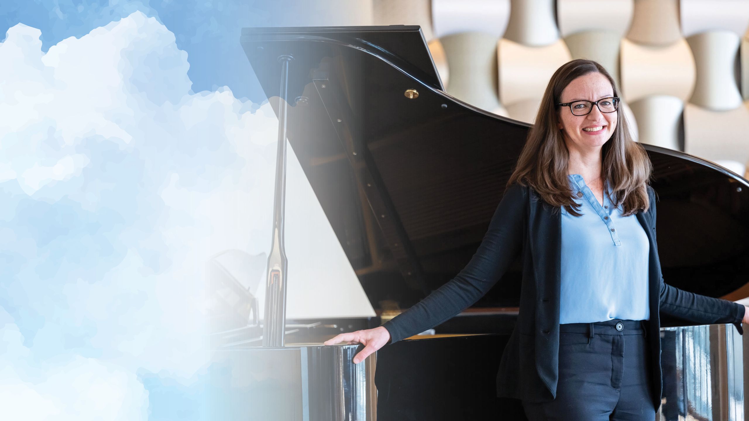 A photo of a white woman with glasses standing in front of a piano and smiling.