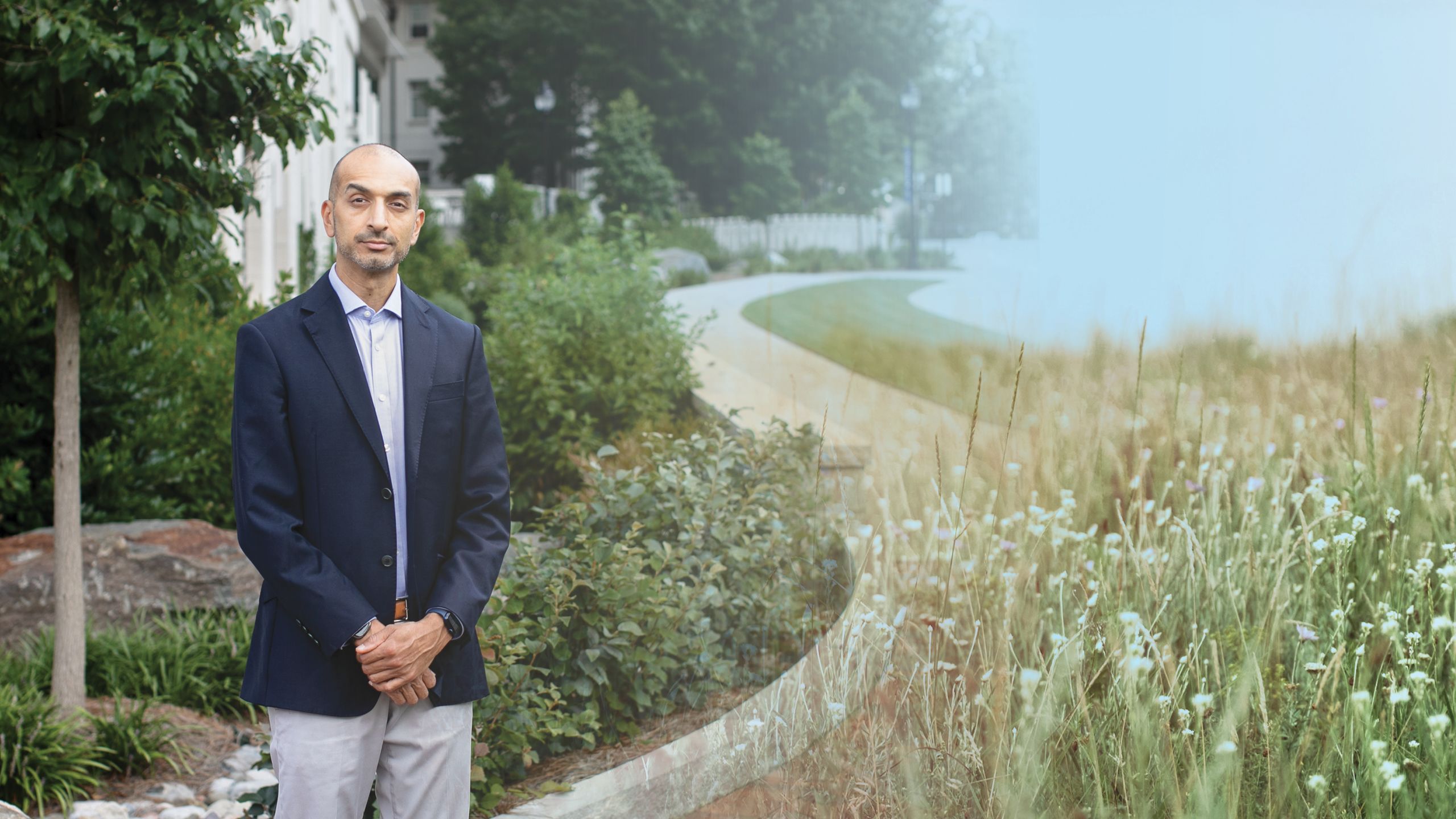 a photo illustration background of a wildflower field on the right and amit shah on the left in front of a building with trees with his hands folded in front of him.