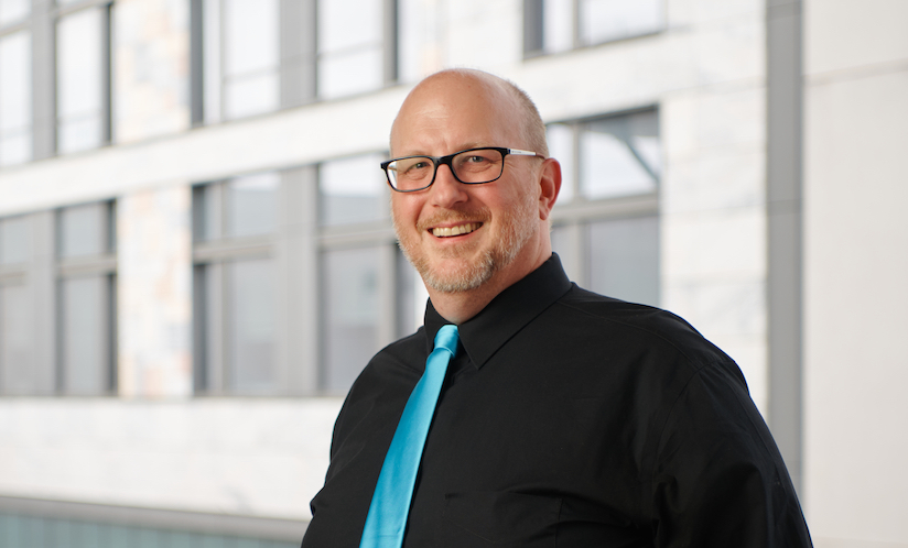 Smiling man with black suit, teal tie, and glasses