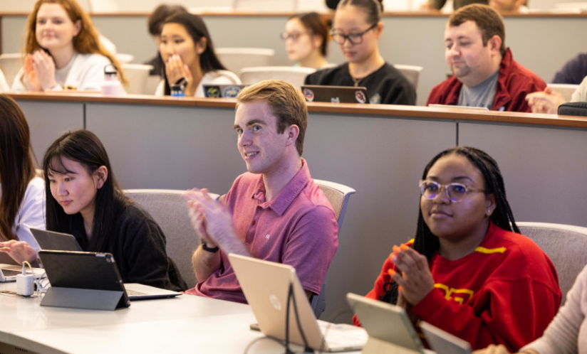 A group of students sit in two rows at lecture hall desks with laptops