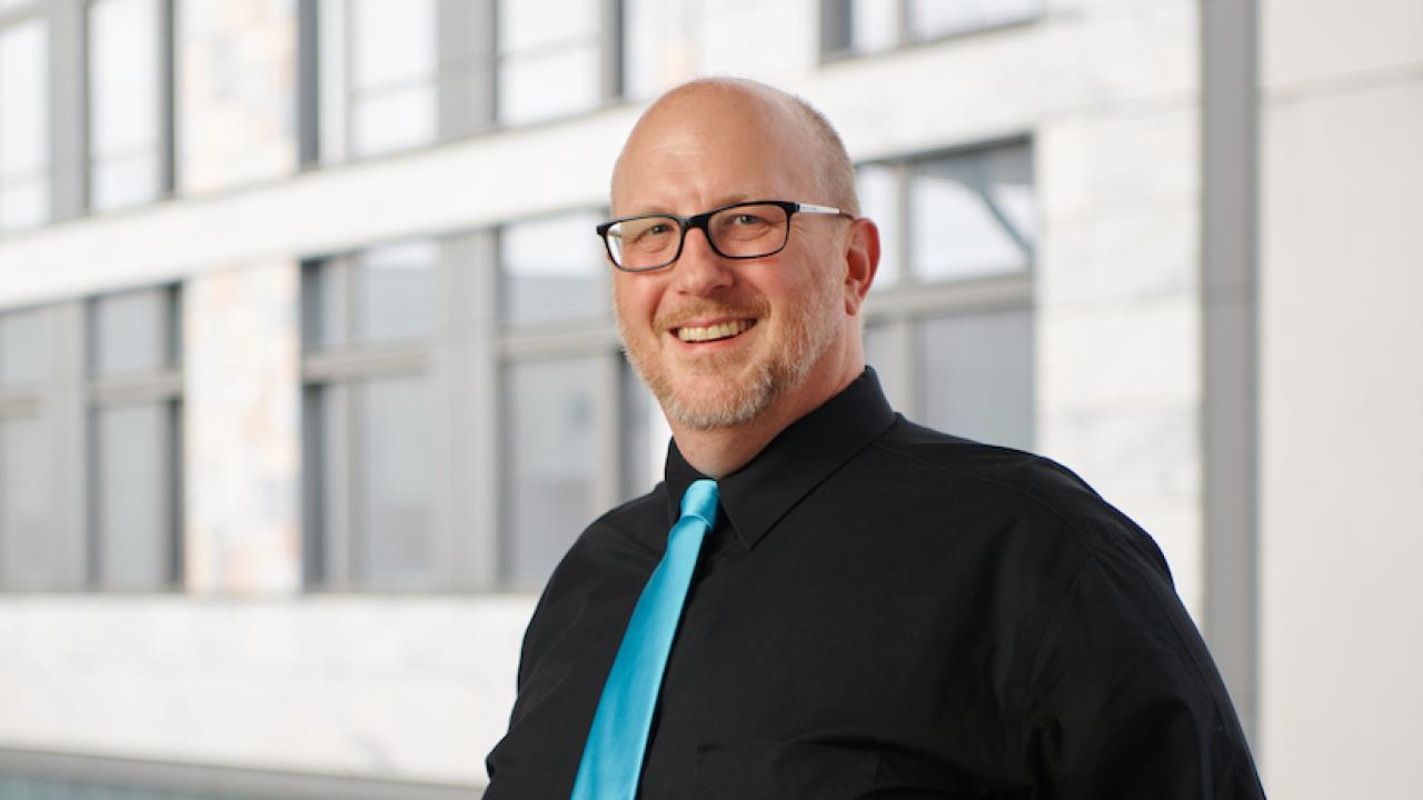 Smiling man with black suit, teal tie, and glasses