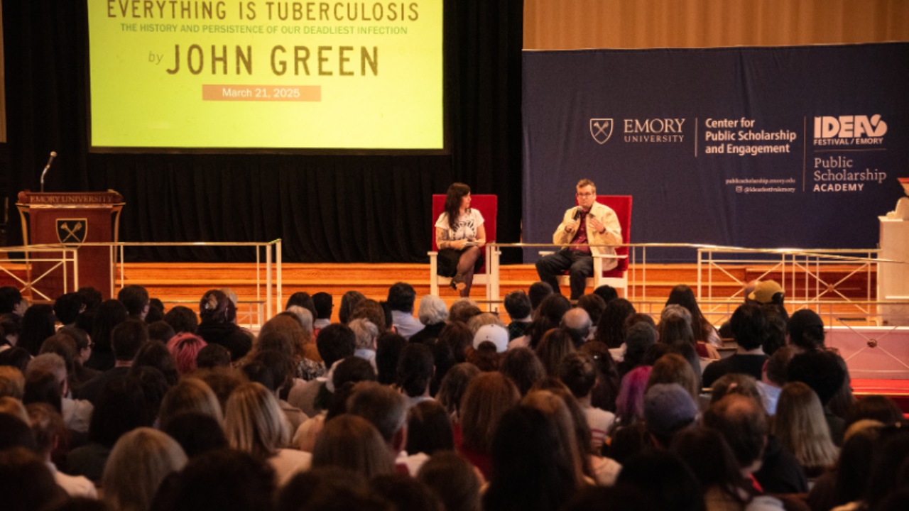 Two people sit on a stage in front of a yellow screen that reads, "Everything is Tuberculosis by John Green"