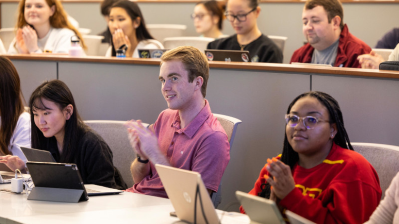 A group of students sit in two rows at lecture hall desks with laptops